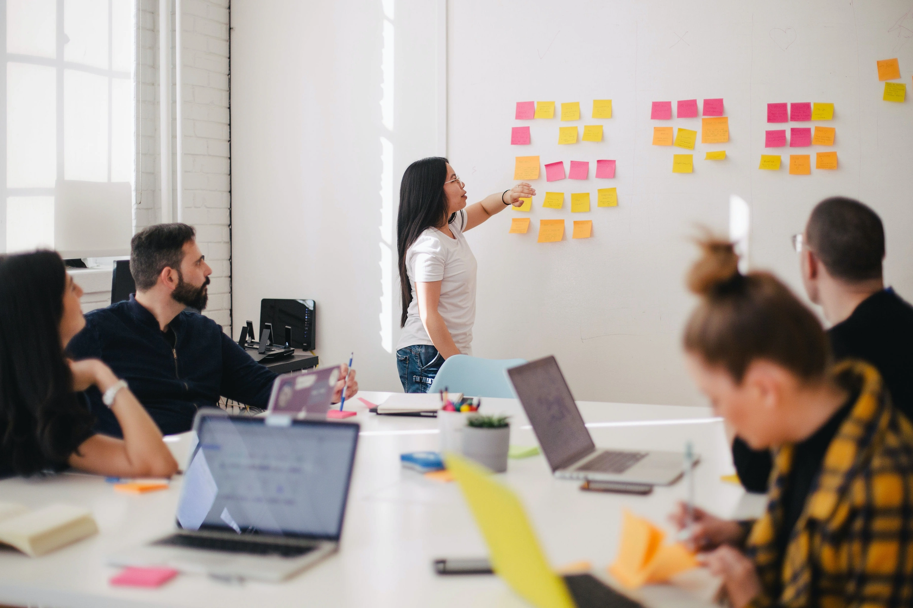 A team meeting around a table with laptops while a woman arranges sticky notes on the wall