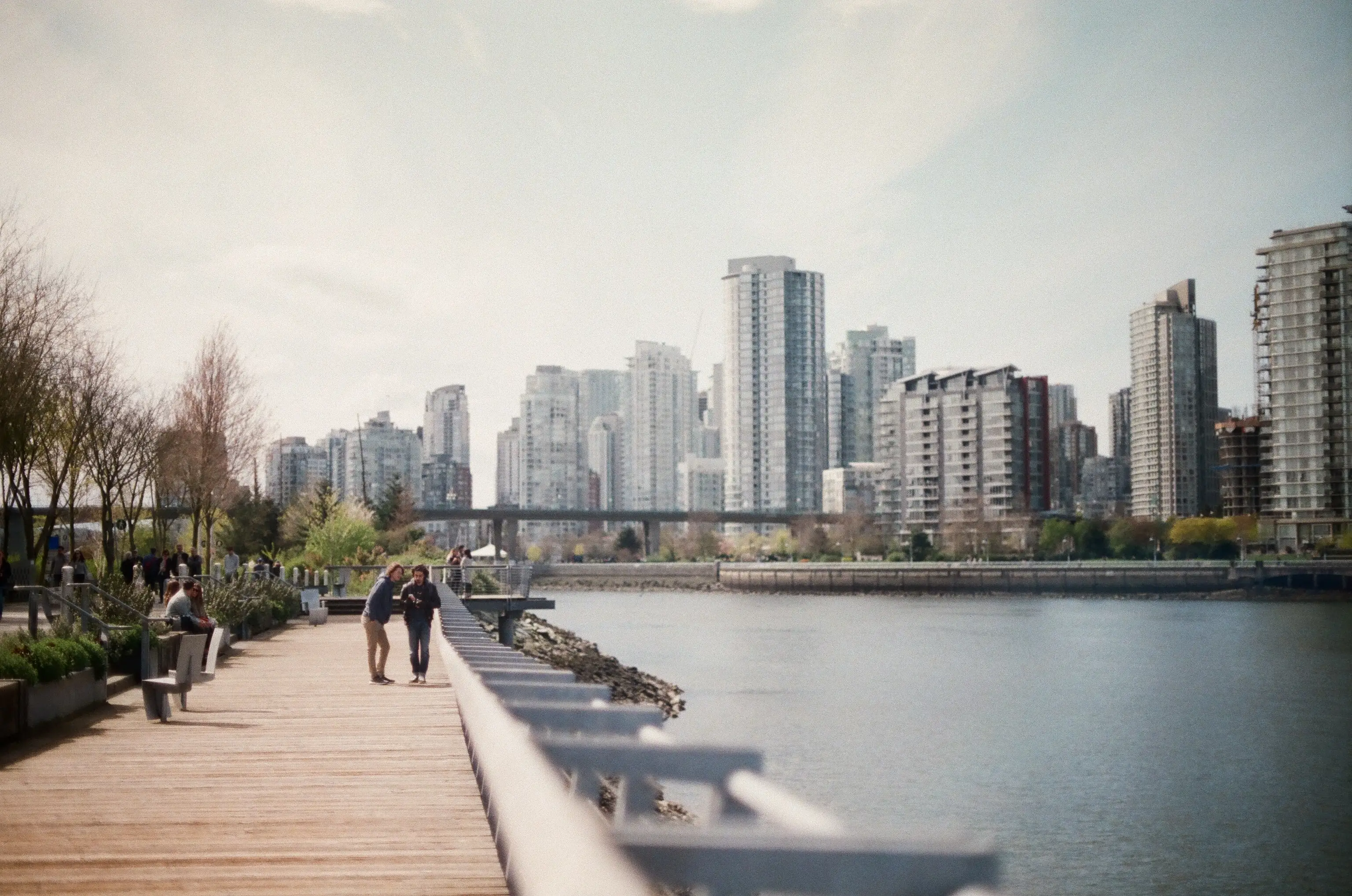 People walking along a waterfront boardwalk beside a city skyline of high-rise towers