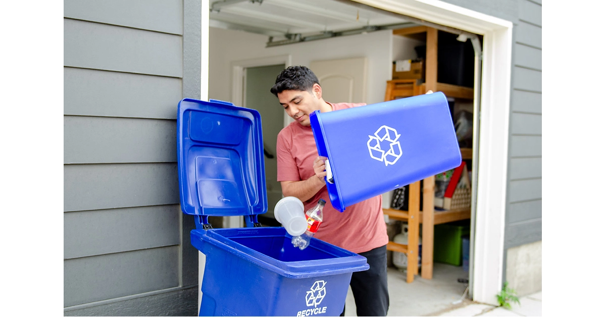 A man pouring a bottle into a blue recycling cart outside his garage