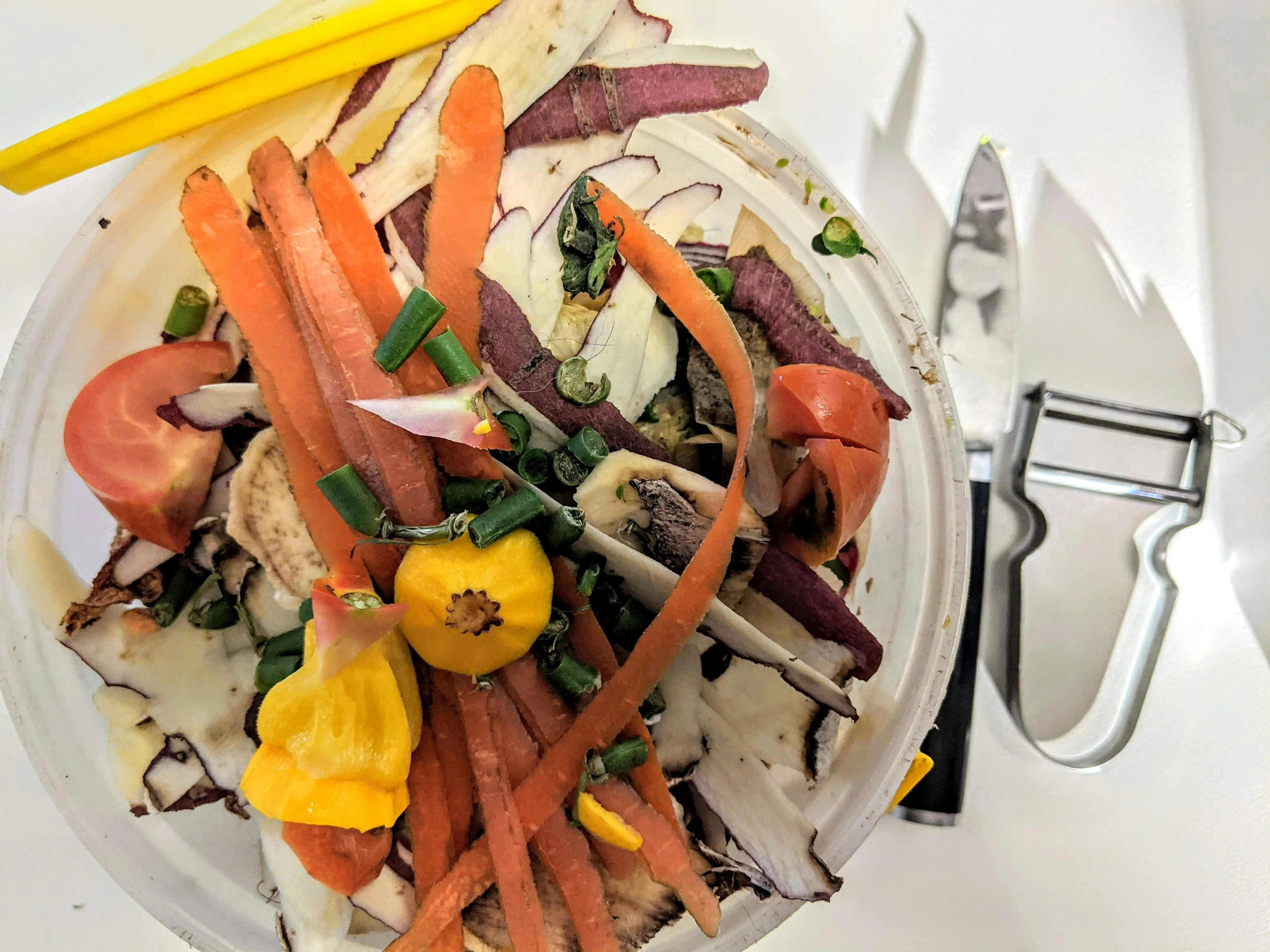 Vegetable peels and food scraps in a compost container beside a peeler