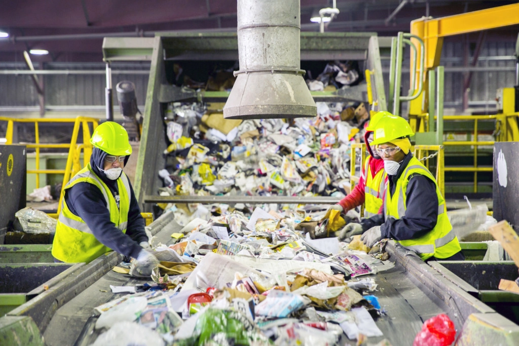 Workers in hard hats and safety vests sorting paper on a recycling facility conveyor