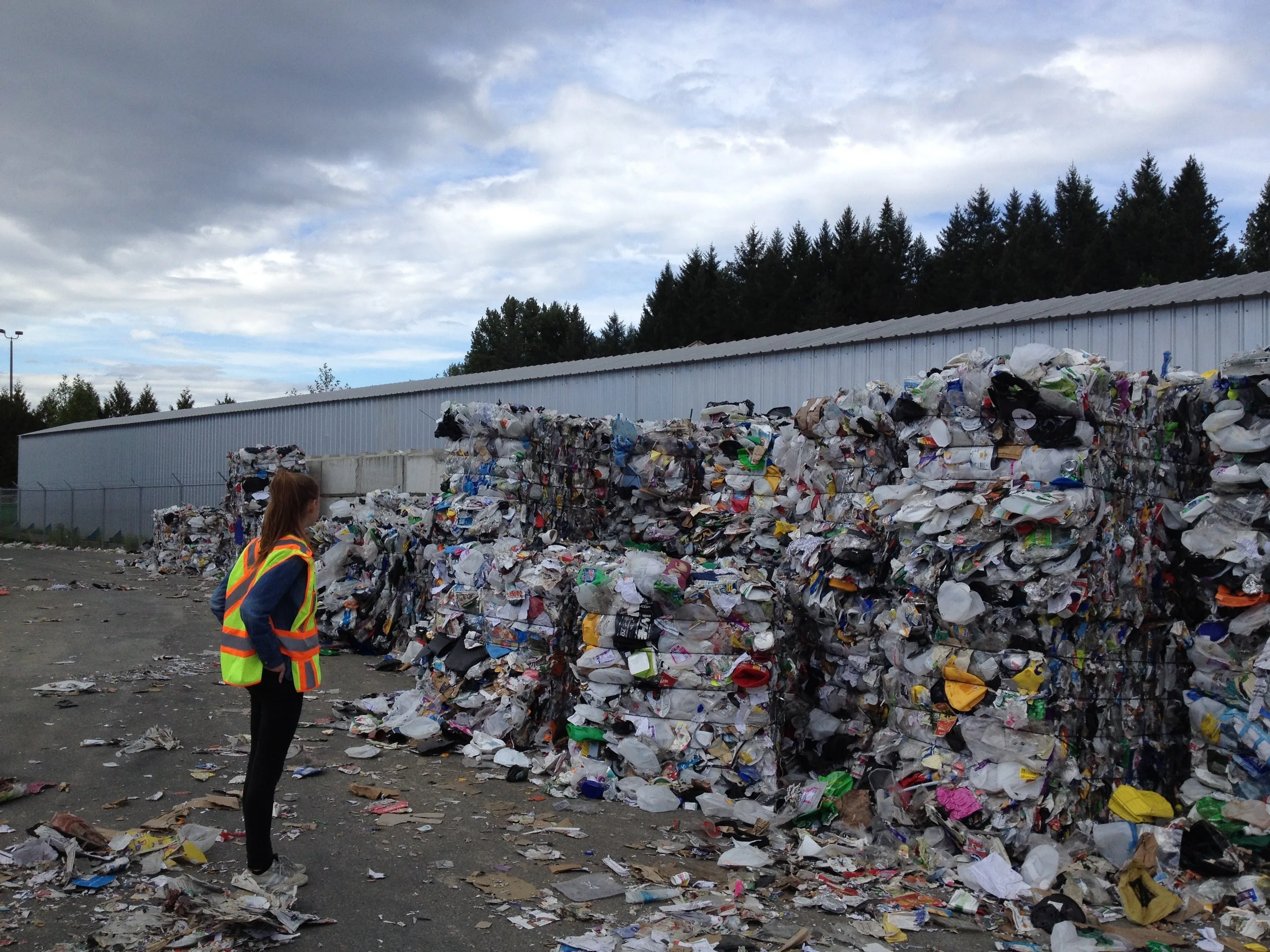 A woman in a safety vest standing beside stacked bales of mixed recyclables outside a facility