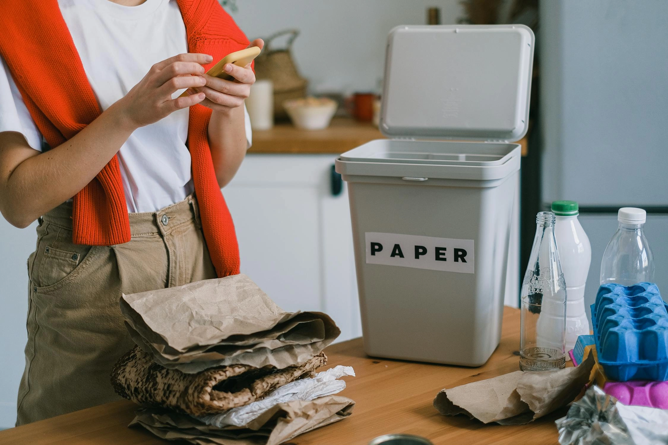 A woman holding a phone beside a paper recycling bin, with sorted paper and plastic bottles on the counter