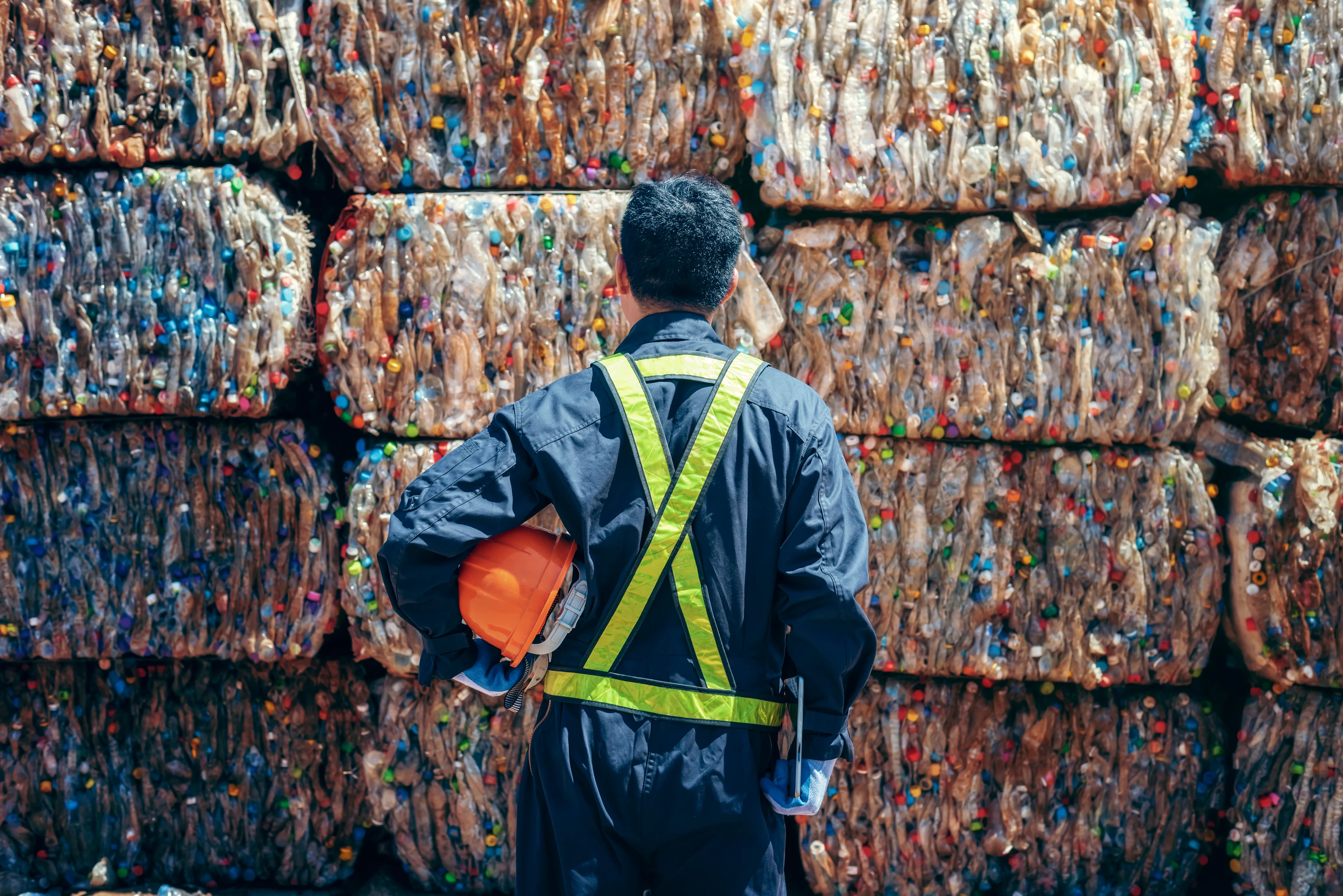 A worker in a safety vest holding a hard hat in front of stacked bales of plastic bottles
